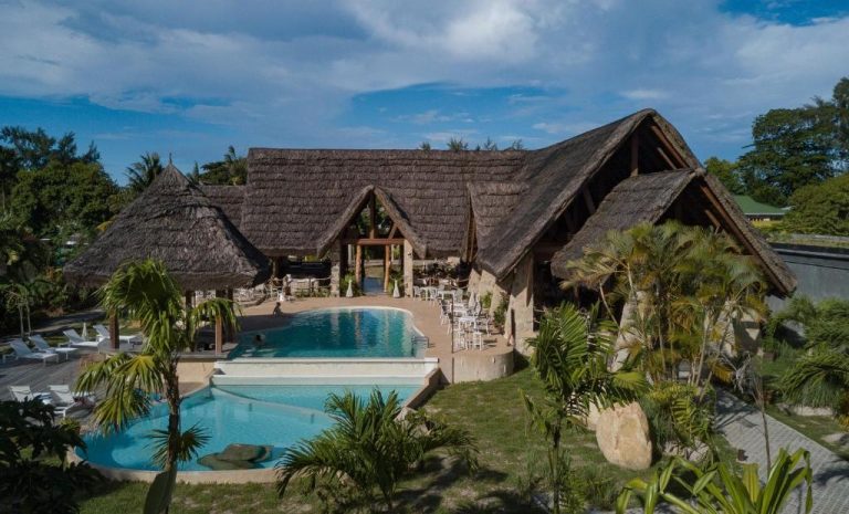 Piscina exterior rodeada de palmeras y restaurante con techo de paja en Les Lauriers Eco Hotel