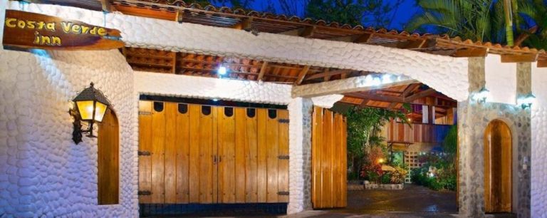 Entrada principal del Costa Verde Inn iluminada de noche con puertas de madera y muros de piedra blanca