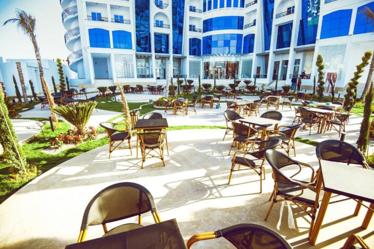 Terraza exterior con mesas y sillas del Lamar Azur Hotel, rodeada de jardines y fachada blanca con cristales azules