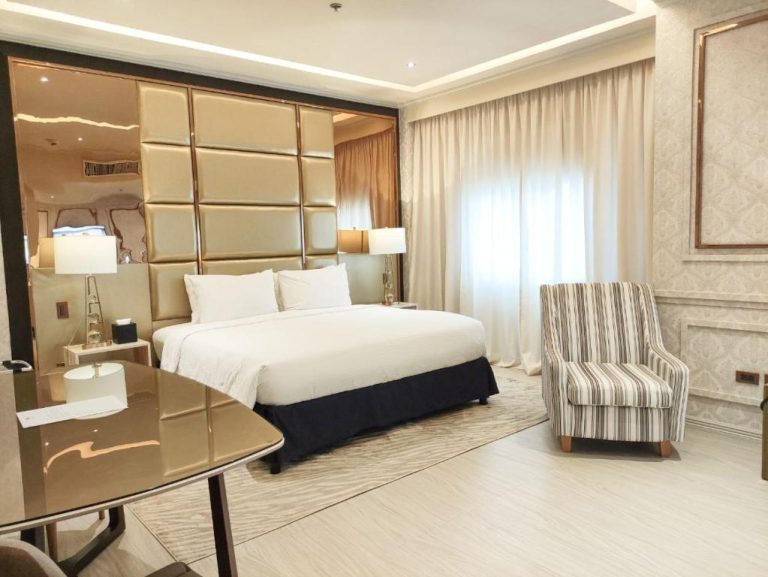 Habitación deluxe con cama king size y cabecero dorado acolchado en The Monarch Hotel