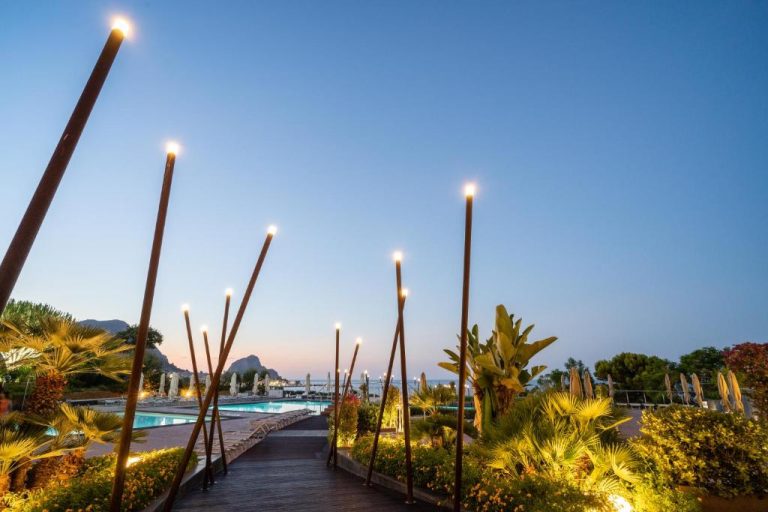 Piscina exterior con jardines tropicales iluminados al atardecer en Domina Zagarella Sicily