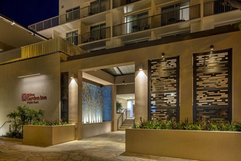 Entrada exterior iluminada del Hilton Garden Inn Waikiki Beach con fuente decorativa y jardines tropicales