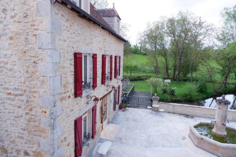 Fachada de piedra con contraventanas rojas y fuente del Moulin de Petoulle rodeado de jardín
