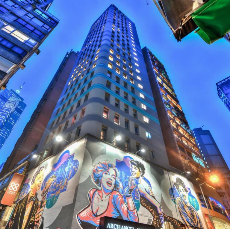 Fachada exterior del Hotel Madera Hollywood con murales de arte urbano y rascacielos iluminado de noche