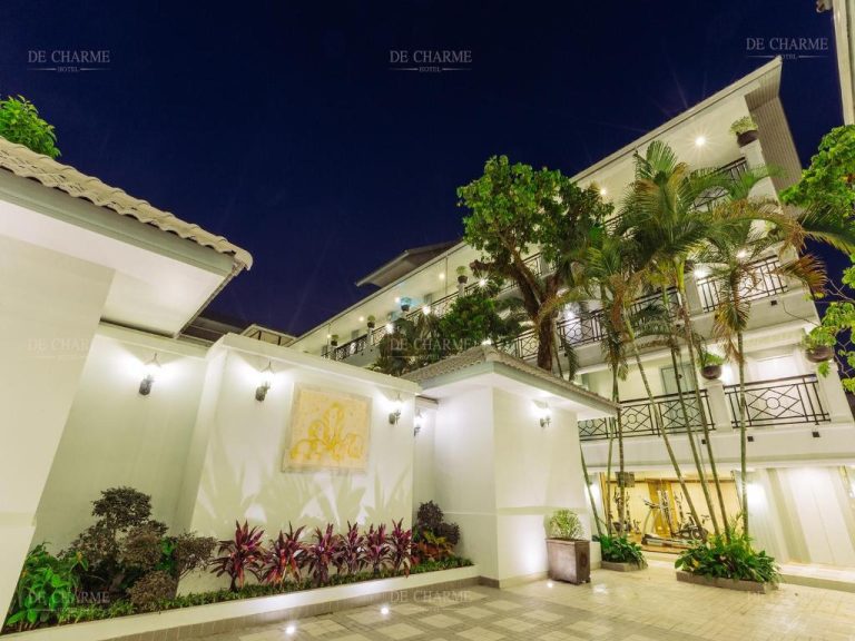 Fachada exterior iluminada del Decharme Hotel con jardines tropicales y palmeras durante la noche