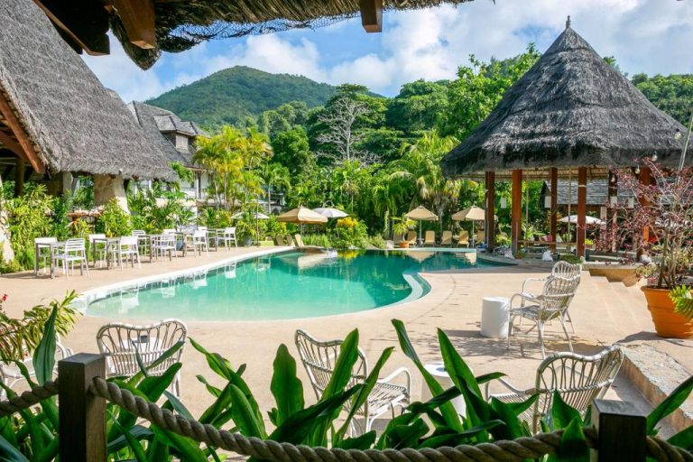 Piscina exterior rodeada de vegetación tropical en Les Lauriers Eco Hotel con cabañas de techo de paja