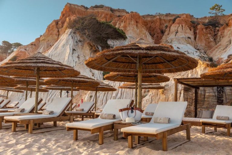 Tumbonas con sombrillas de paja en la playa del EPIC SANA Algarve Hotel con acantilados rojos al fondo