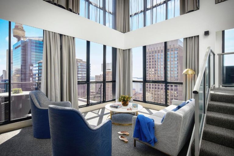 Salón luminoso con vistas al skyline de Síndney en Meriton Suites Pitt Street con sillones azules