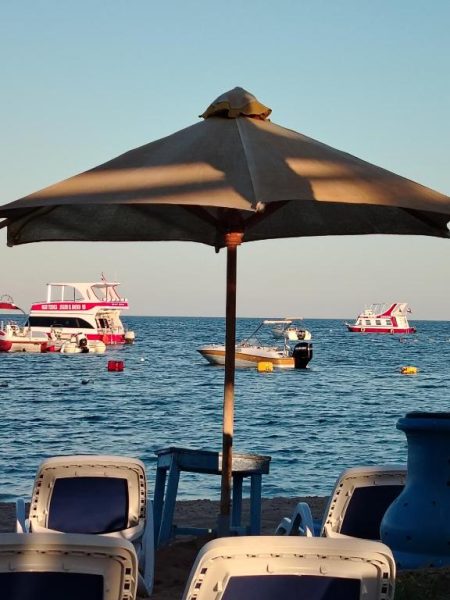 Tumbonas y sombrilla en la playa del Hotel Kanabesh con barcos anclados en el mar al fondo