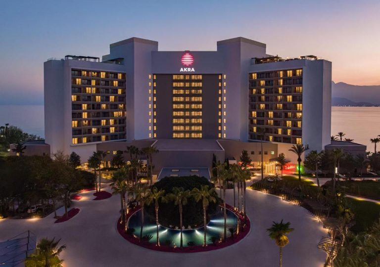 Vista aérea de la fachada exterior del Hotel Akra Antalya iluminada al atardecer con jardines y fuente