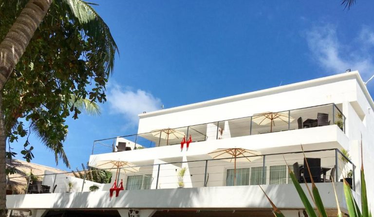 Fachada exterior del Sundown Beach Studios con terrazas, sombrillas y tumbonas rojas bajo cielo azul