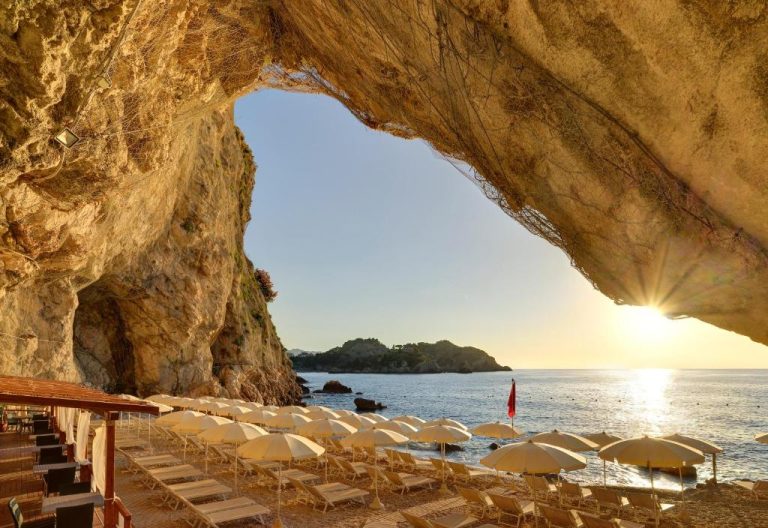 Playa privada con tumbonas y sombrillas del UNA Hotels Capotaormina vista desde una cueva rocosa al atardecer