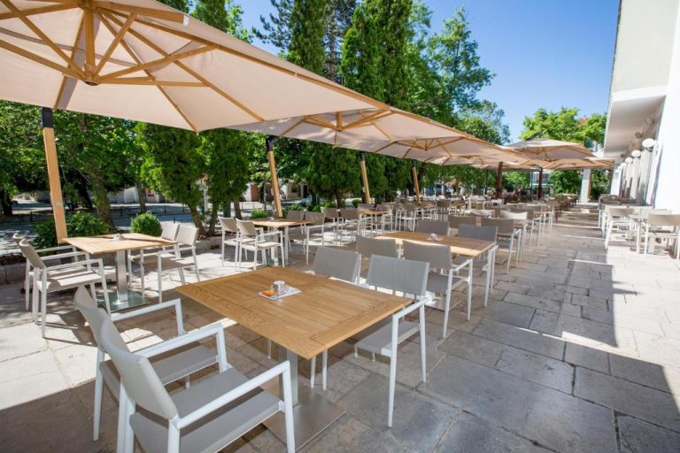 Terraza exterior del Hotel Onogošt con mesas de madera, sillas blancas y grandes sombrillas beige rodeada de árboles