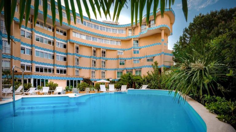 Piscina exterior con tumbonas y fachada curva del Savoia Hotel Rimini rodeada de palmeras