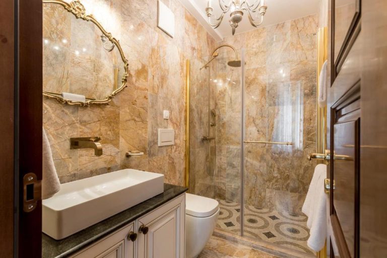 Baño de lujo con mármol dorado, ducha de cristal y espejo clásico en Villa Nepos Hotel