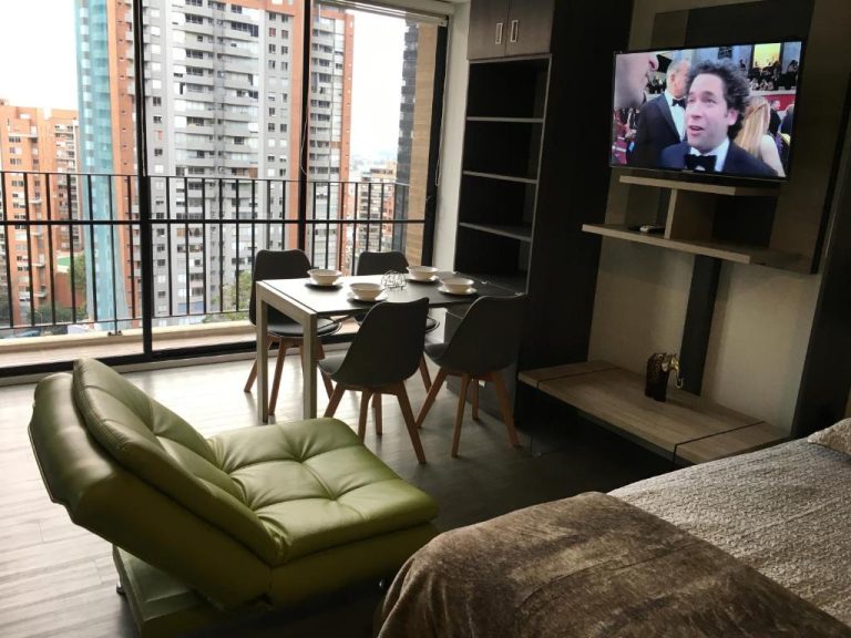 Estudio moderno con sala comedor, silla verde, televisión y vista panorámica a la ciudad en Enjoy In Bogotá