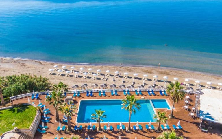 Vista aérea de la piscina exterior y playa privada del Crystal Beach Hotel con tumbonas y sombrillas