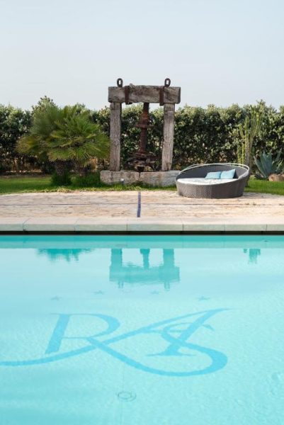 Piscina exterior con logo del Relais Antiche Saline, zona de relax y antiguo lagar en el jardín