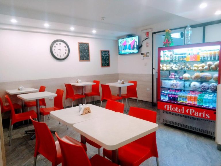 Cafetería del Hotel París Lima con mesas blancas, sillas rojas y vitrina de alimentos iluminada