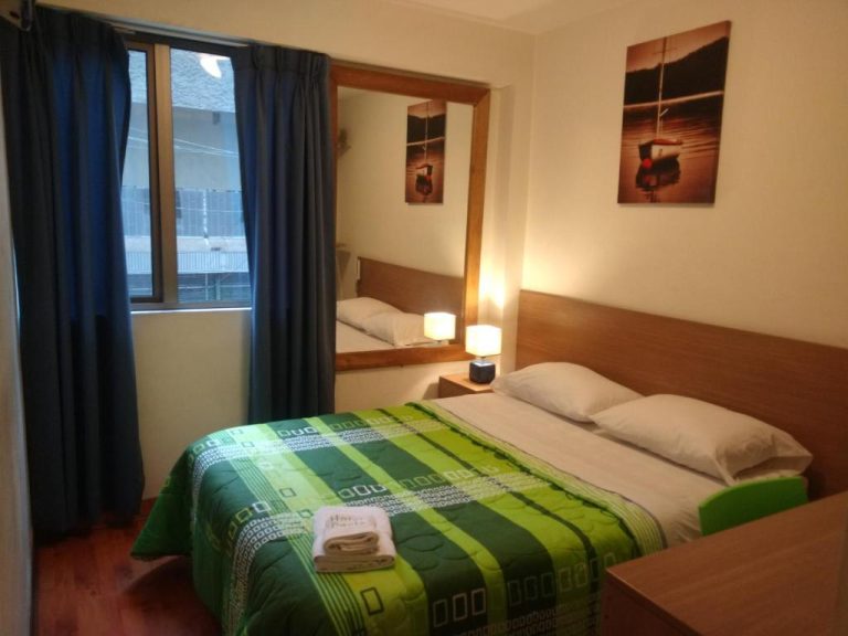 Habitación doble con cama matrimonial y colcha verde en Hotel Paris Lima, espejo y ventana con cortinas azules