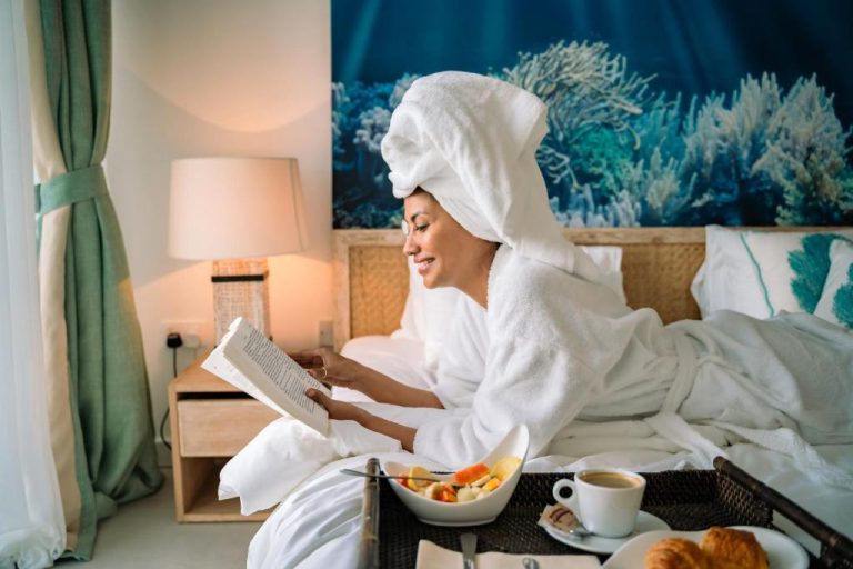 Mujer en bata blanca disfrutando del desayuno en cama con libro en suite del AQUA Boutique Hotel
