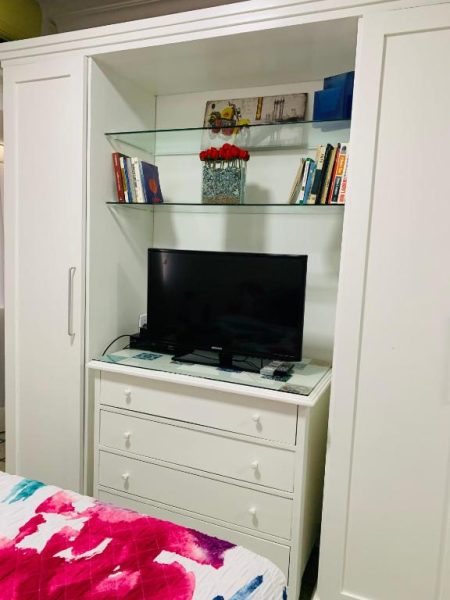 Habitación del Studio Cristo Carnaval con televisor Samsung, cómoda blanca y estantes con libros y flores rojas