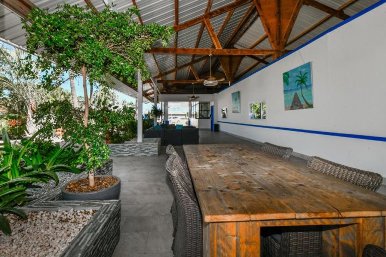 Terraza comedor exterior con mesa de madera y jardín tropical en Aruba Blue Village Hotel and Apartments