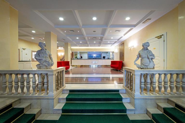 Lujoso lobby del Ambassador Palace Hotel con estatuas clásicas, escalinata de mármol y recepción iluminada