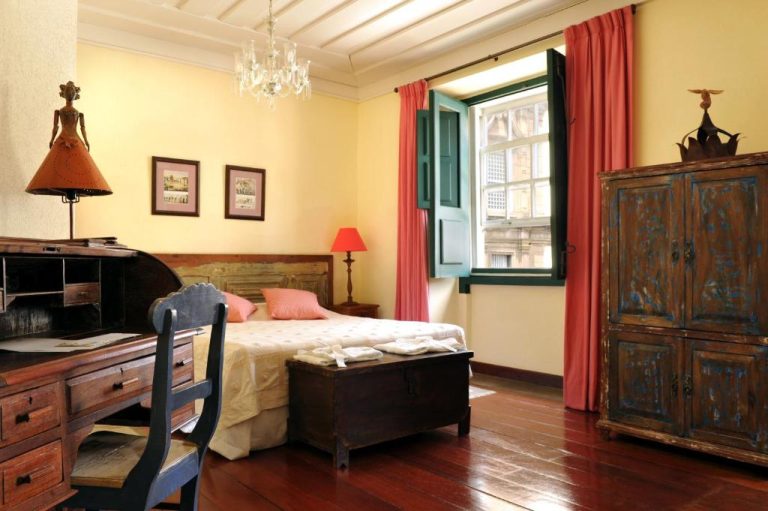 Habitación colonial con muebles antiguos de madera y ventana con cortinas rojas en Hotel Villa Bahia