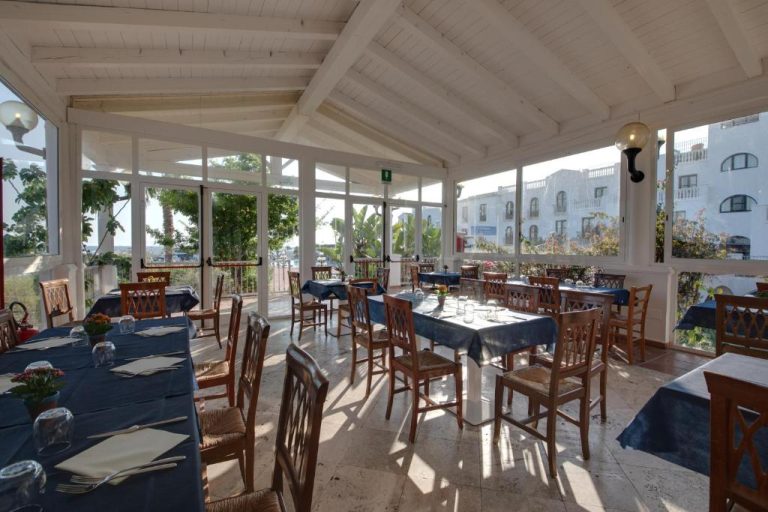 Restaurante con terraza acristalada y mesas con mantel azul en Phi Hotel Sighientu, Cerdeña