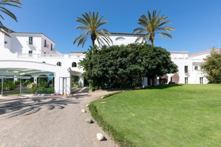 Fachada blanca y jardines exteriores del Phi Hotel Sighientu rodeado de palmeras en Cerdeña