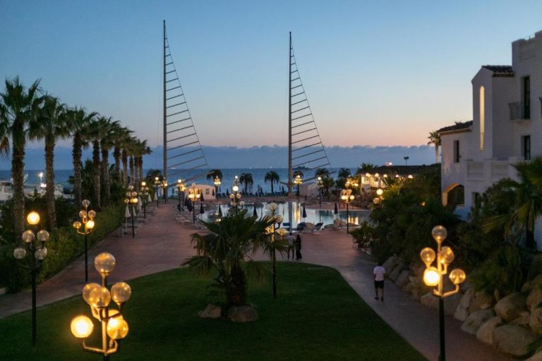 Piscina exterior al atardecer con palmeras y paseo iluminado del Phi Hotel Sighientu frente al mar