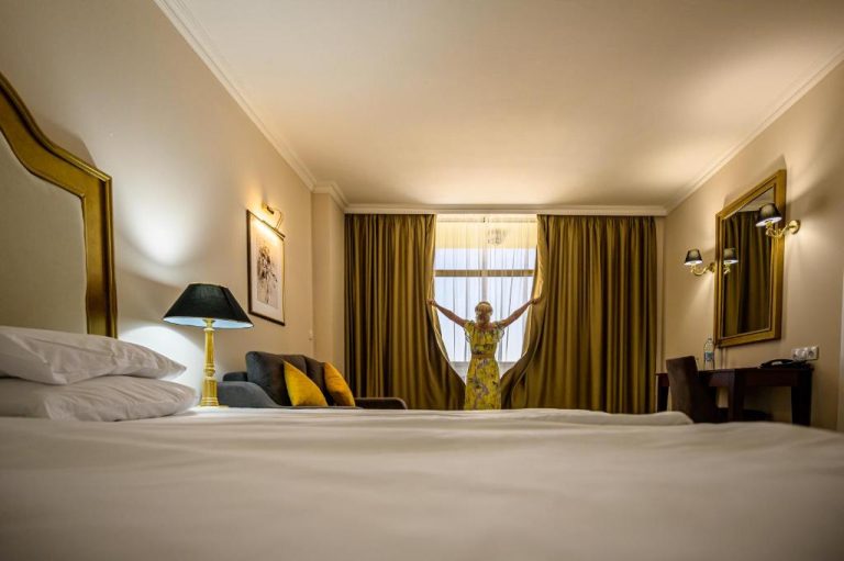 Mujer abriendo cortinas doradas en habitación de lujo del Imperial Palace Hotel con cama king size