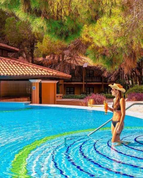 Mujer disfrutando de la piscina exterior del Asteria Family Resort Belek rodeada de pinos