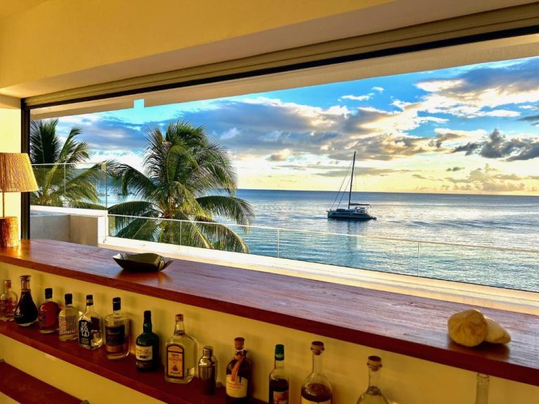 Bar con vista panorámica al mar y velero en La Plage Martinique con palmeras y atardecer