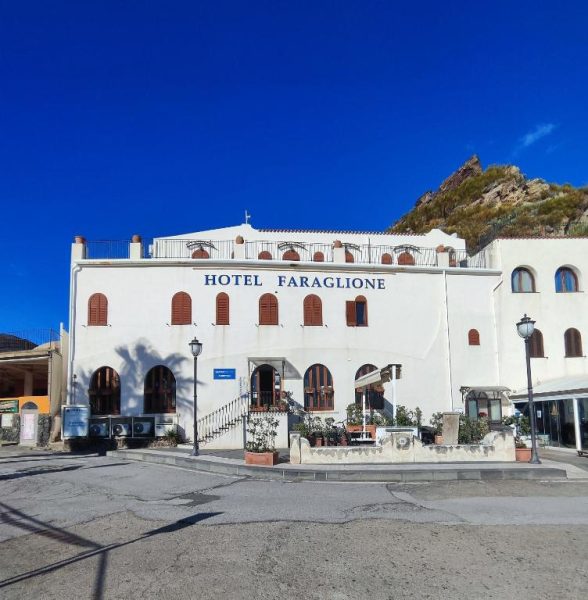 Fachada exterior blanca del Hotel Faraglione con montaña rocosa al fondo y cielo azul