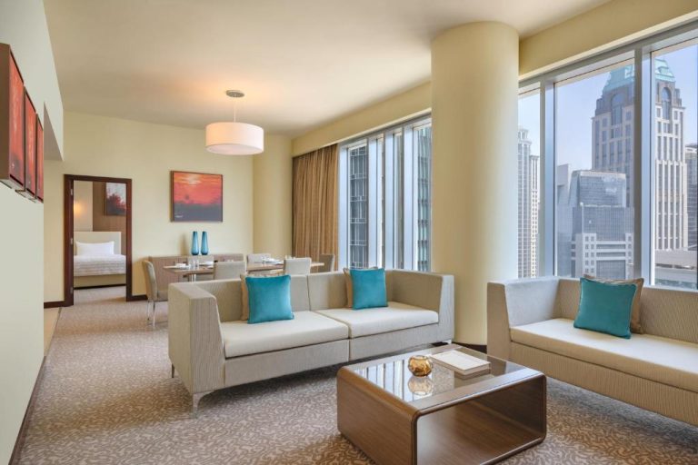 Salón de suite con sofás blancos y cojines turquesa con vistas al skyline en Le Meridien City Center Doha