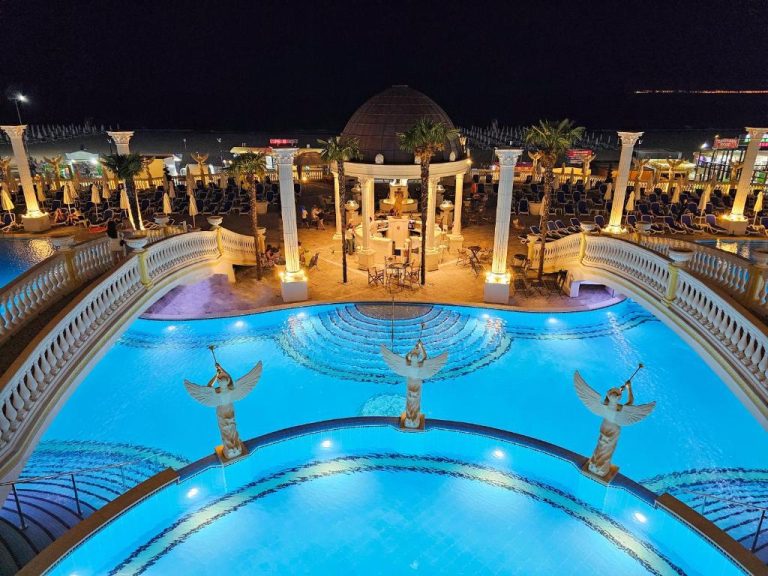 Piscina iluminada de noche con estatuas de ángeles y gazebo clásico en Imperial Palace Hotel