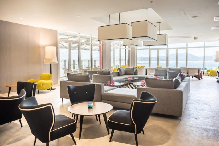 Amplio salón lounge del Simon Hotel con sofás grises, sillones amarillos y vistas panorámicas al mar