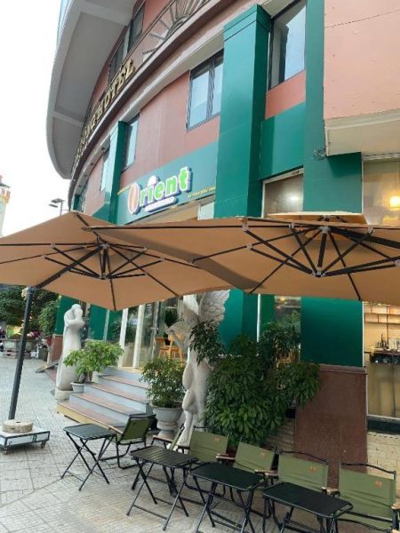 Terraza exterior con mesas, sillas y parasoles del Orient Hotel con fachada verde y esculturas decorativas