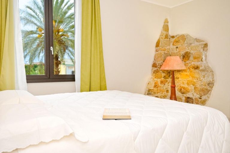 Habitación doble con cama blanca y pared de piedra decorativa en Hotel Corallo New con vistas a palmeras