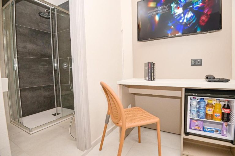 Habitación con ducha de cristal, escritorio y minibar con bebidas en Hotel Corallo New