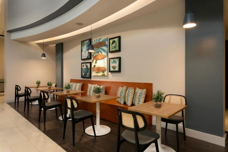 Área de comedor con mesas de madera y sofá del Hyatt Place Manatí, decorada con cuadros y plantas