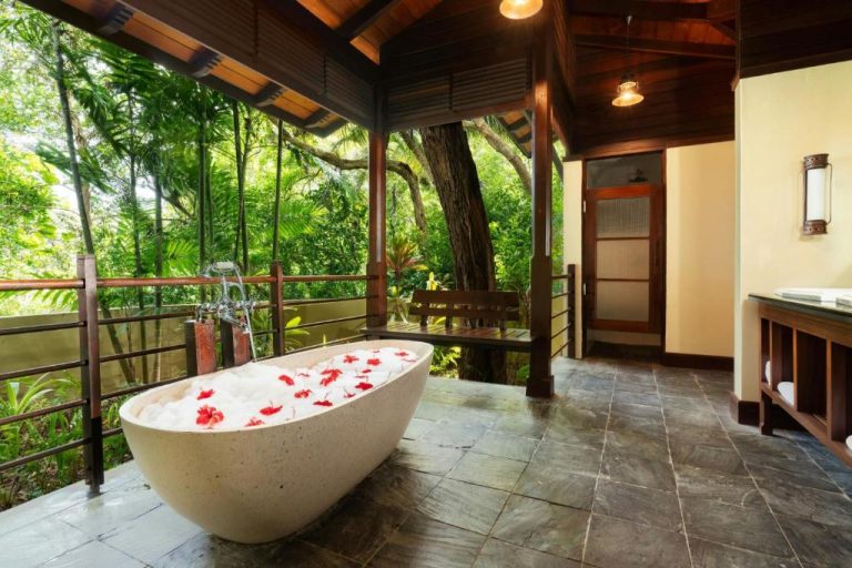 Bañera exenta de piedra con pétalos de flores en terraza abierta con vistas a la selva en Enchanted Island Resort