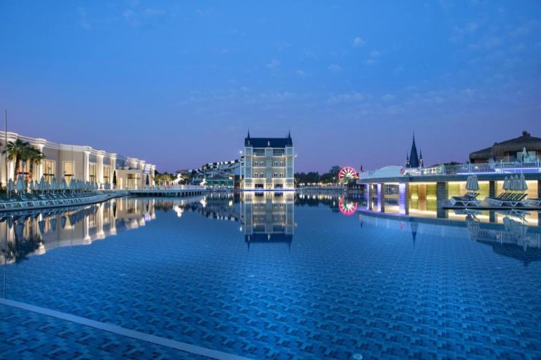 Piscina principal iluminada de noche en Granada Luxury Belek con reflejo del edificio central