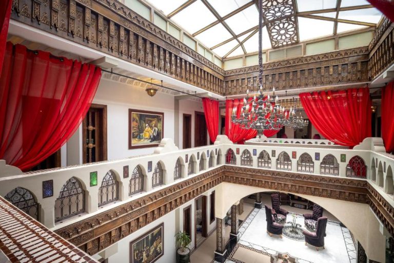 Patio interior de lujo con arcos árabes, lámpara de araña y cortinas rojas en Hotel Ryad Art Place Marrakech