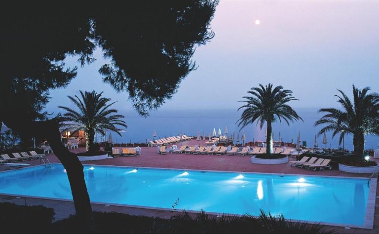 Piscina exterior iluminada de noche con vistas al mar en el Grand Hotel Baia Verde
